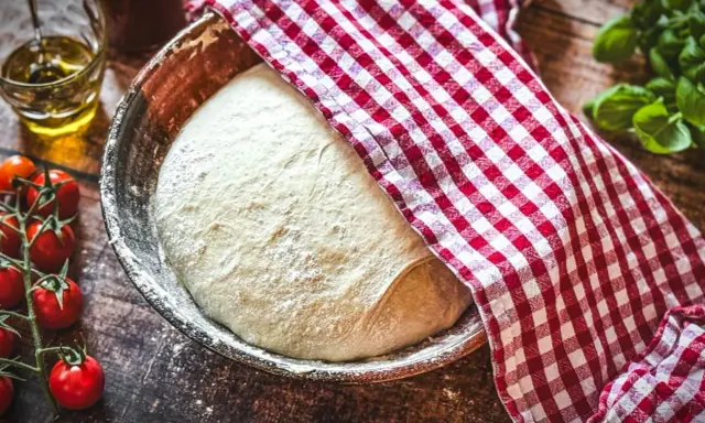 Pizzateig original rezept von einem italienischen gastronomen ruht in einer Schüssel, bedeckt mit einem rot-weißen Tuch. Tomaten und Olivenöl daneben.