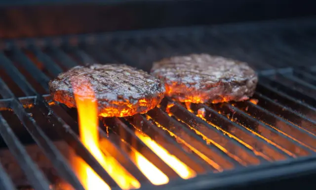 Zwei Burger-Patties auf einem Gasgrill, die Flammen lecken an ihnen hoch. Die richtige Temperatur ist entscheidend für das perfekte Burger-Grillen.