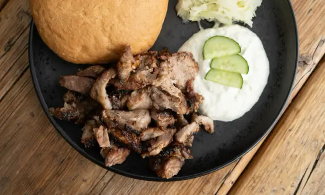 Saftiges Gyros vom Grill mit Tzatziki, Krautsalat und einem Brötchen auf schwarzer Platte.
