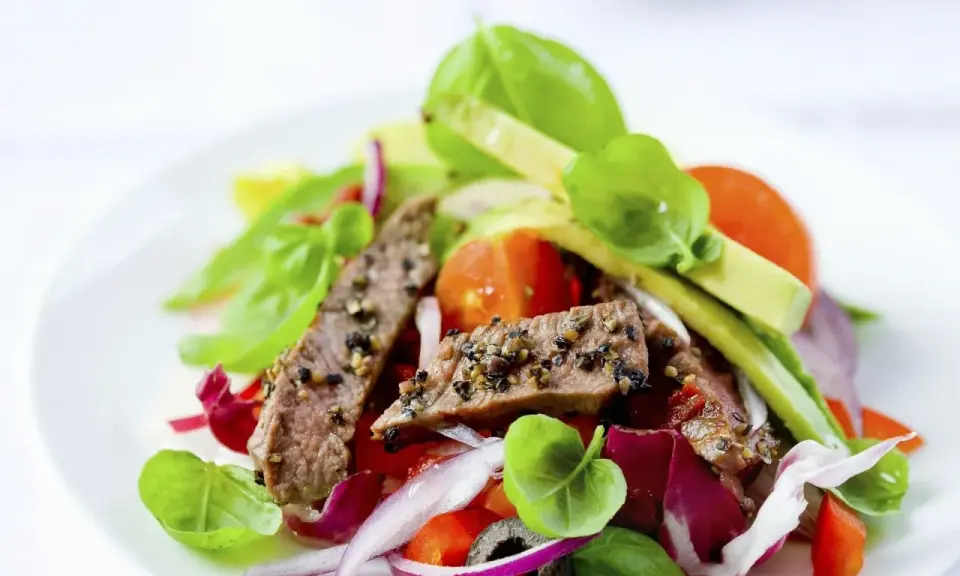 Frischer Salat mit zarten Rinderstreifen, Tomaten, roten Zwiebeln und Basilikum auf einem weißen Teller.