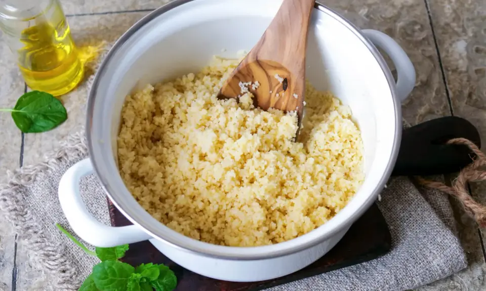 Ein Topf mit lockerem Couscous, bereit für ein marokkanisches Rezept. Ein Holzlöffel rührt die Körner auf. Daneben eine Flasche Öl und frische Minze.