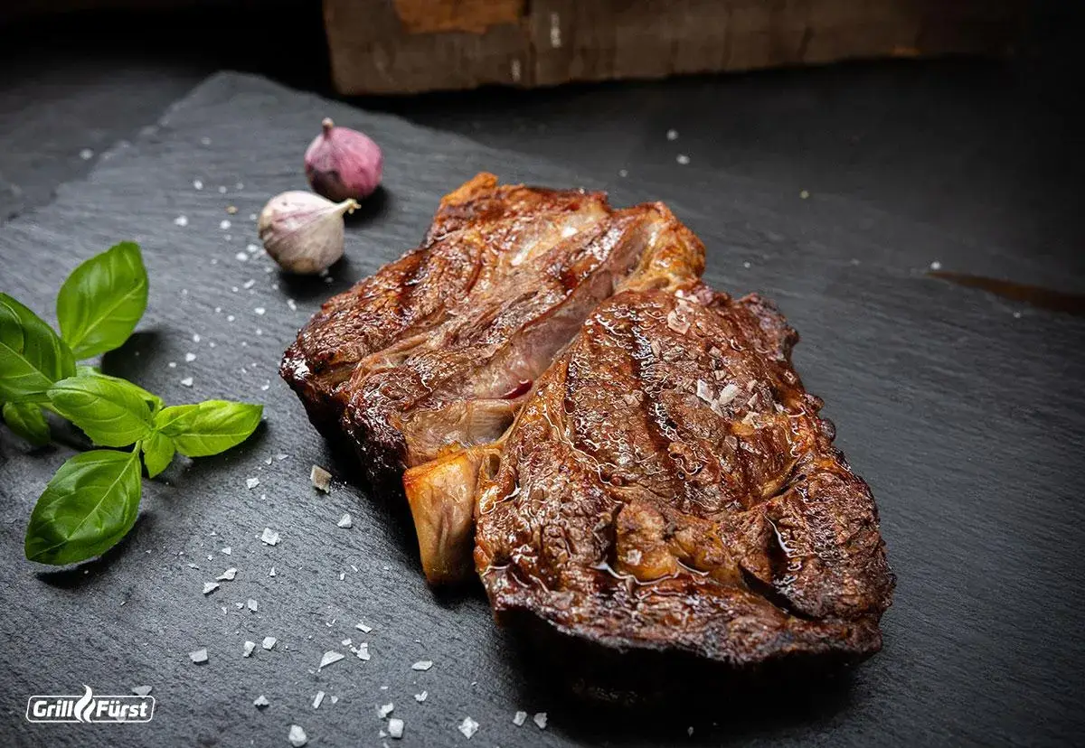 Perfekt gegrilltes Rib Eye Steak mit grobem Salz, Basilikum und Knoblauch auf schwarzem Stein.