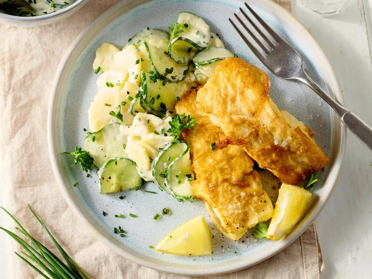 Knuspriger Backfisch mit cremigem Kartoffelsalat und Gurkenscheiben, garniert mit frischer Petersilie und Zitronenspalten.