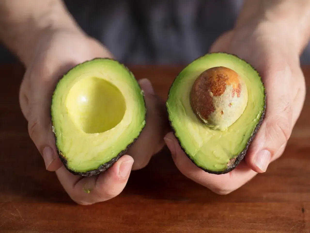 Zwei H&auml;lften einer Avocado in H&auml;nden. Eine ist leer, die andere mit Kern. Geduld ist gefragt, um die Avocado reifen lassen.