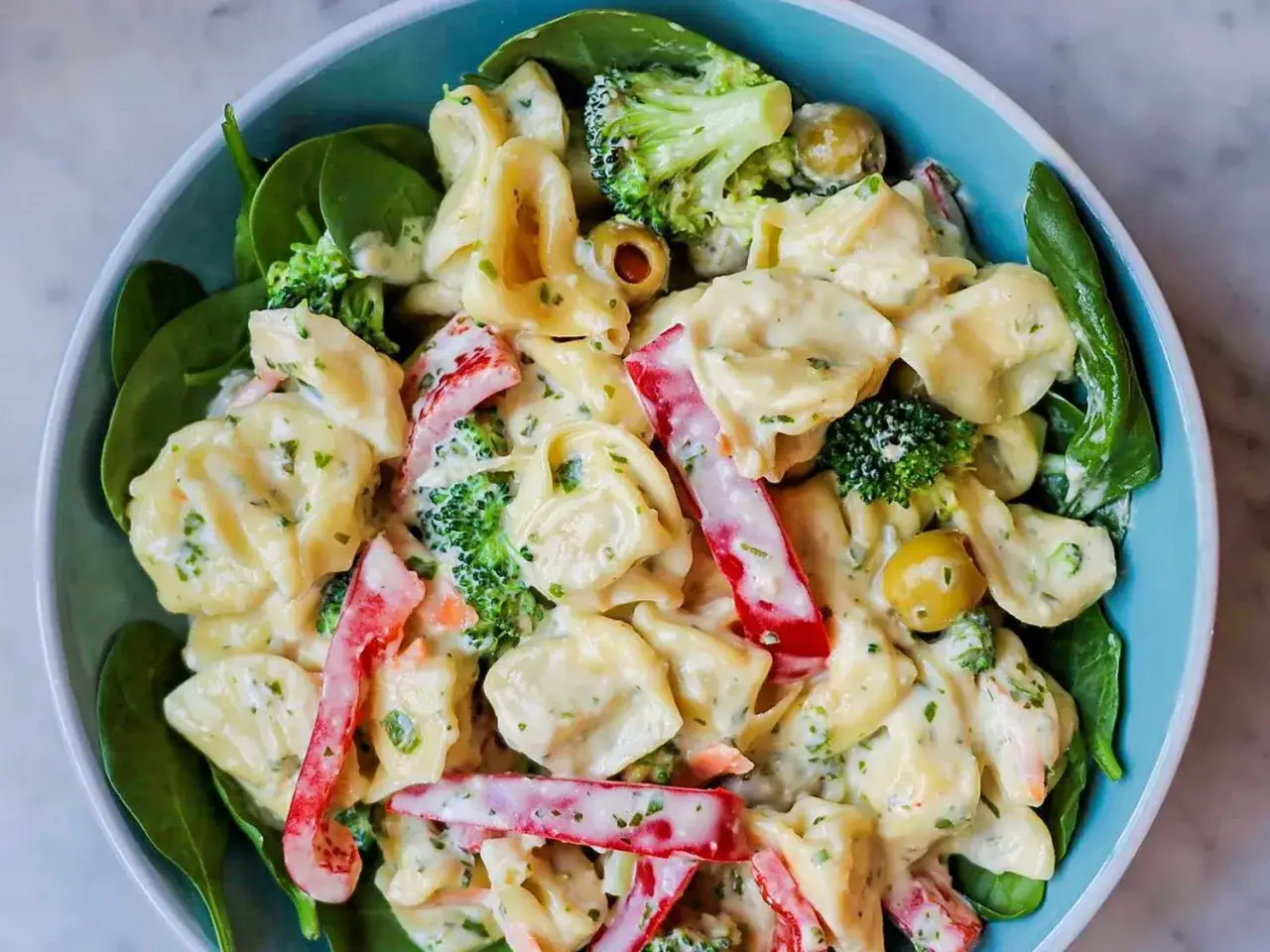 Ein cremiger tortellini salat mit pesto, garniert mit Brokkoli, Paprika und Oliven, serviert auf Spinat.
