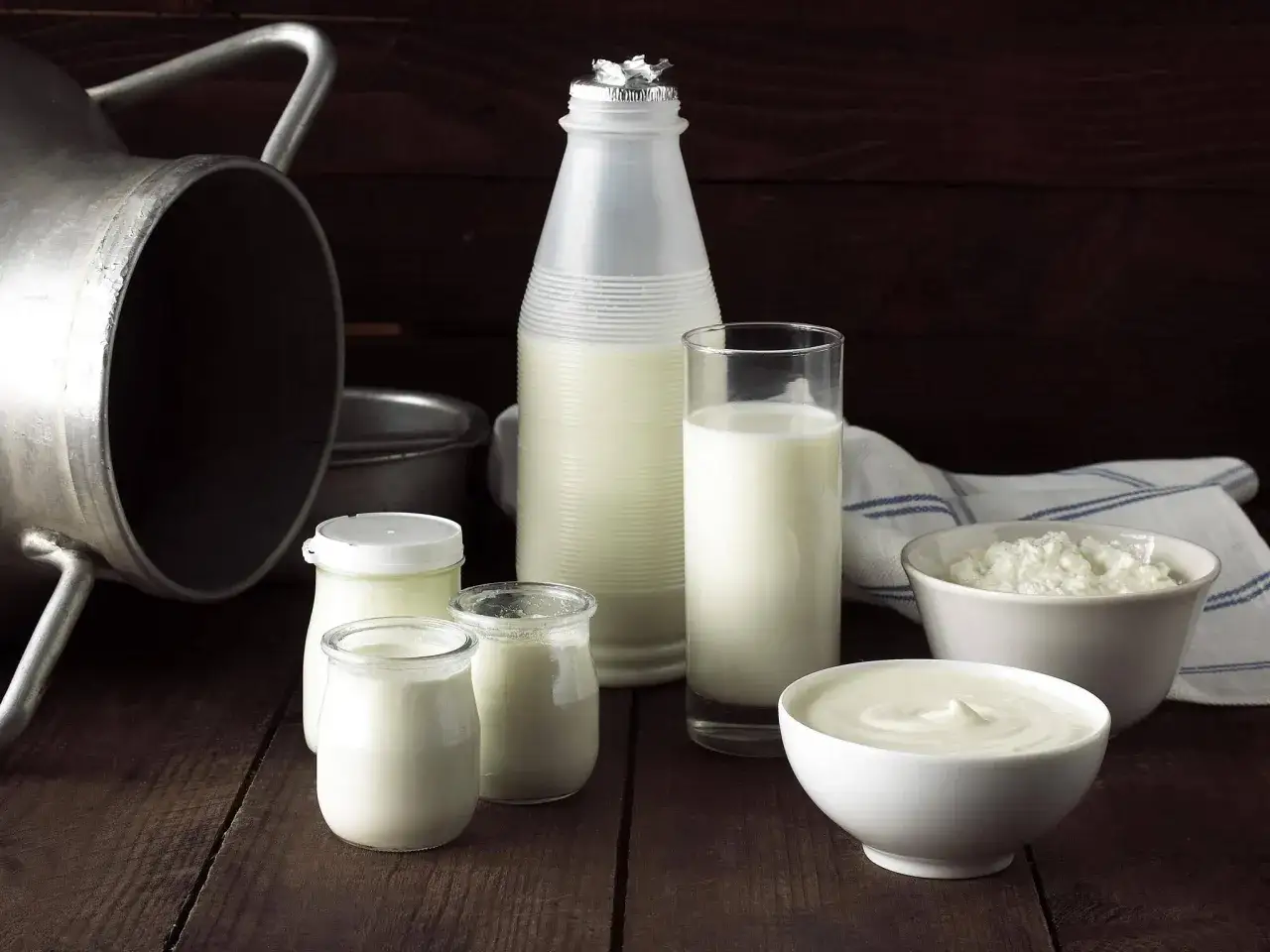 Milchprodukte wie Joghurt und Schmand sind gesund. Ein gro&szlig;er Milchbeh&auml;lter, Flaschen, Gl&auml;ser und Schalen mit Milchprodukten stehen auf einem Holztisch.