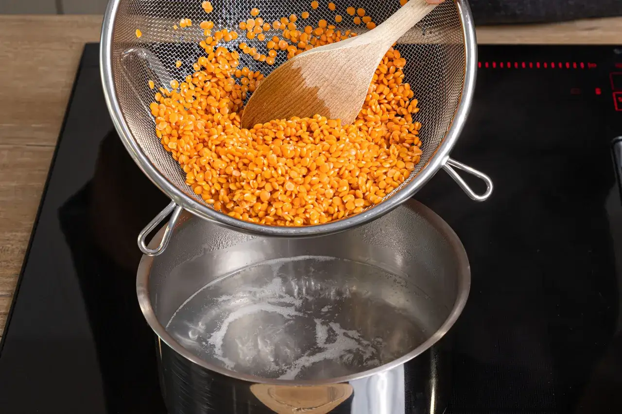 Rote Linsen kochen: Ein Sieb mit roten Linsen wird über einen Topf mit kochendem Wasser gehalten.