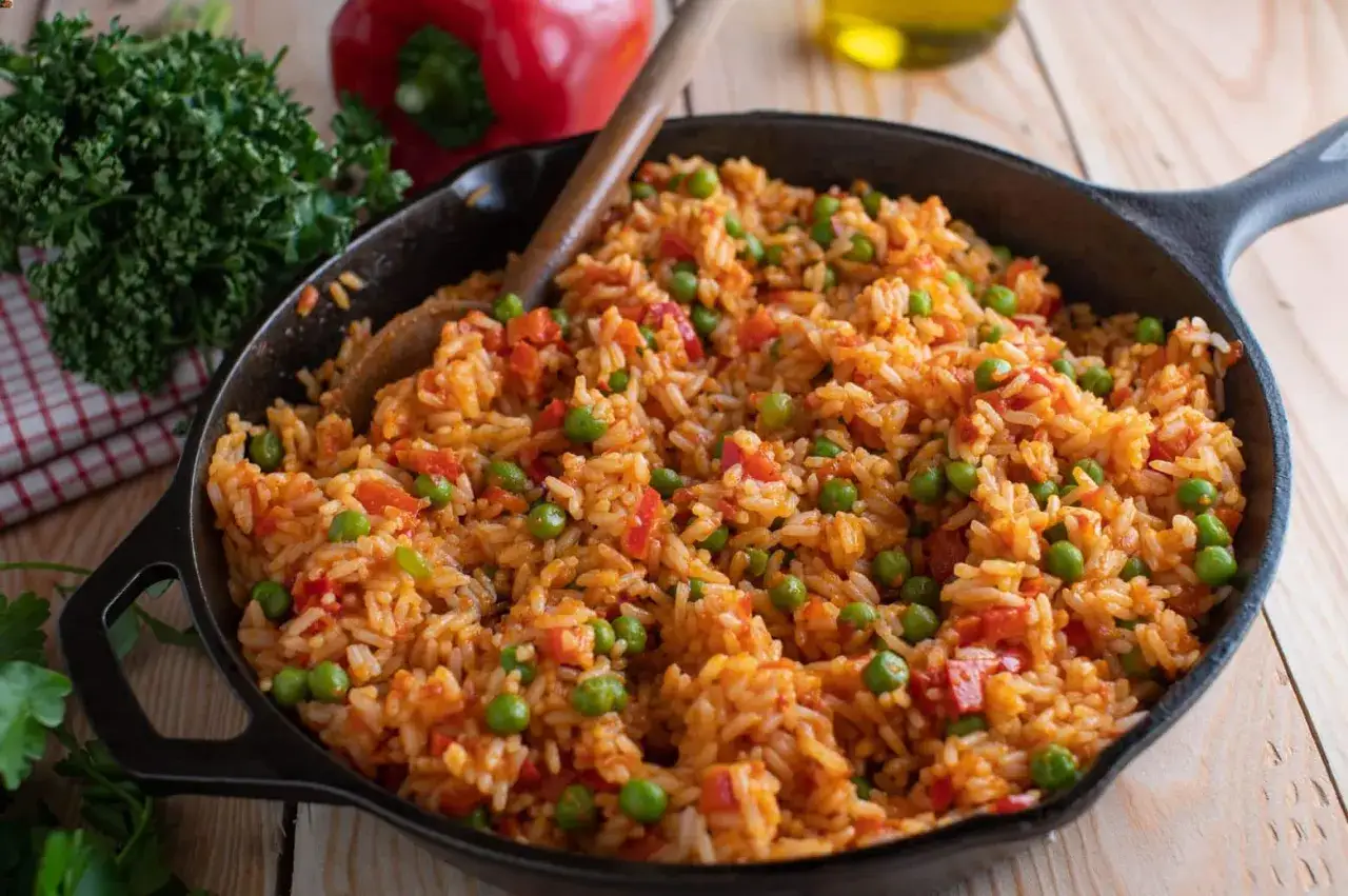 Djuvec Reis in einer Gusseisenpfanne, mit Erbsen und Paprika. Ein Holzkochlöffel liegt darin.