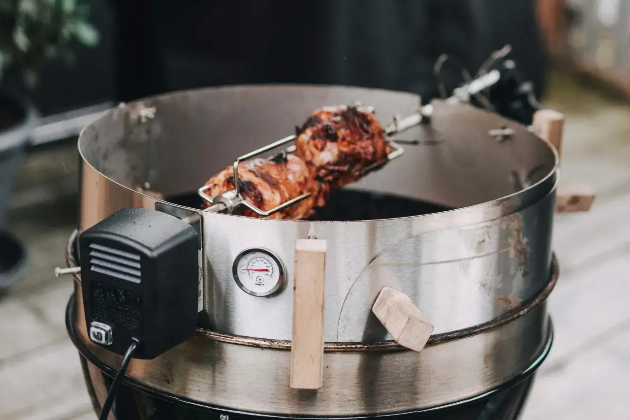 Schweinshaxe sous vide dreht sich am Spie&szlig; &uuml;ber gl&uuml;hender Kohle. Ein Thermometer zeigt die Gartemperatur an.