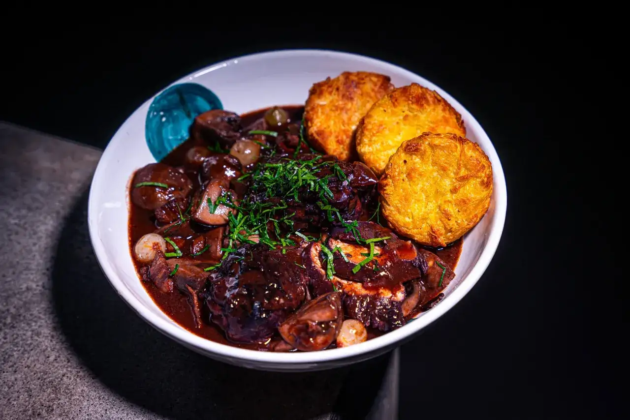 Ein Teller mit Coq au Vin Rezept Original: zartes H&auml;hnchen in Rotwein geschmort, mit Pilzen und Perlzwiebeln, garniert mit frischen Kr&auml;utern und serviert mit knusprigen Kartoffelpuffern.