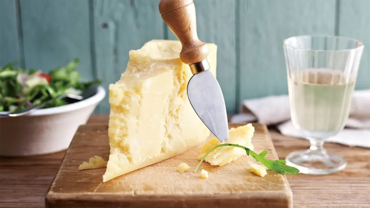 Ein Stück Parmesan wird mit einem Messer auf einem Holzbrett angeschnitten. Daneben ein Glas Weißwein und eine Schüssel Salat. Ist Parmesan vegetarisch?