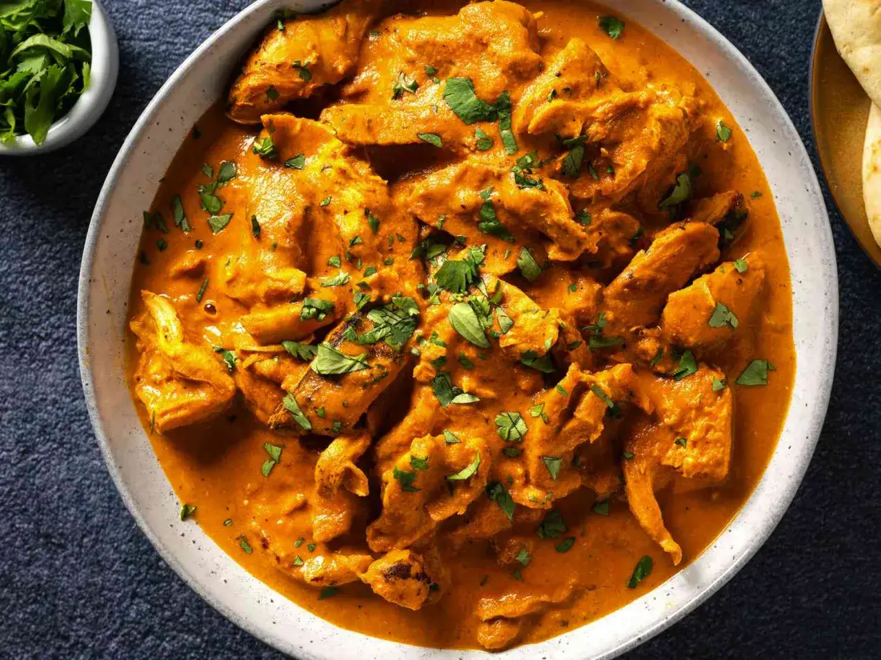 Cremiges Chicken Tikka Masala, garniert mit frischem Koriander, serviert in einer Schüssel. Daneben ein kleiner Napf mit Koriander und ein Stück Naan.