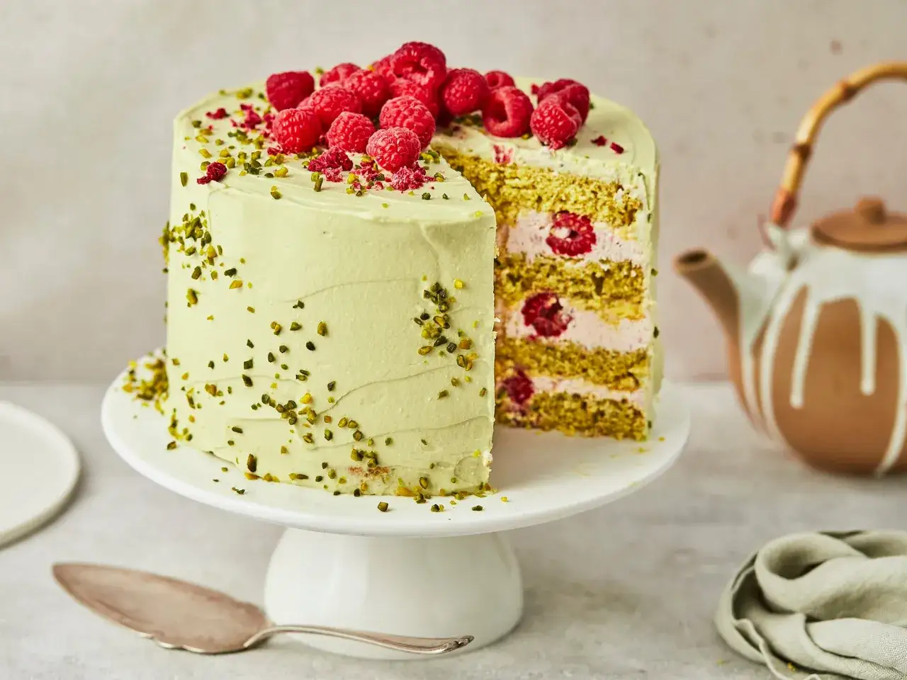 Himbeer-Pistazien-Torte mit zarter gr&uuml;ner Creme, frischen Himbeeren und gehackten Pistazien. Ein St&uuml;ck ist angeschnitten und zeigt die saftigen Schichten.