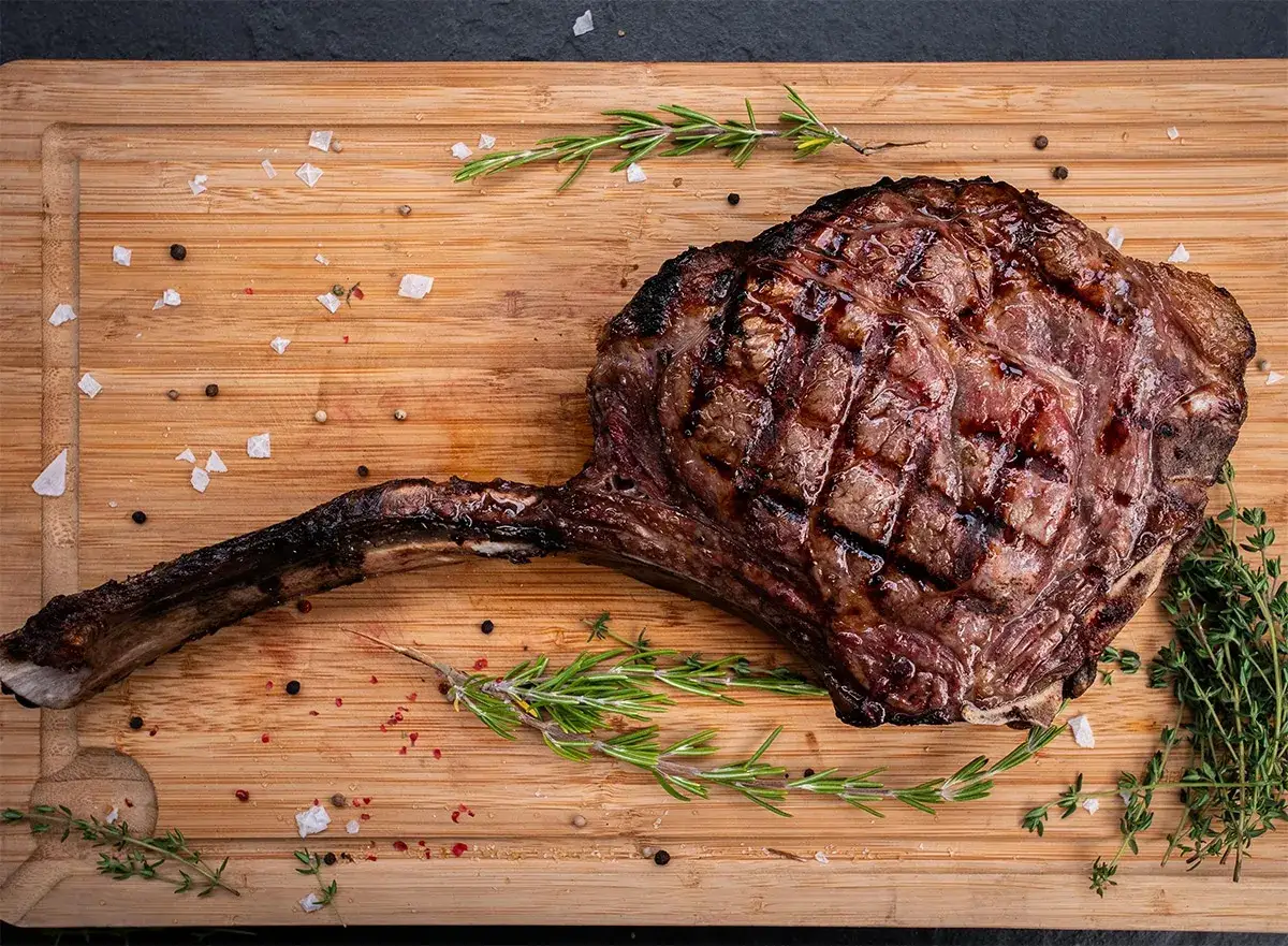 Ein saftiges Tomahawk Steak, perfekt gegrillt, liegt auf einem Holzbrett mit Rosmarin und grobem Salz.