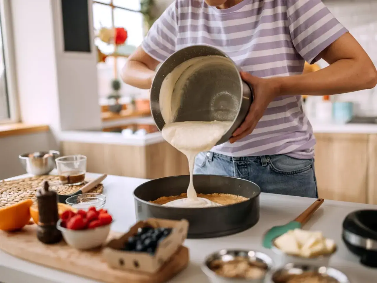 Eine Person gie&szlig;t eine cremige Masse in eine Springform, um einen Kuchen zu backen. Zutaten wie Beeren und Orangen liegen bereit.