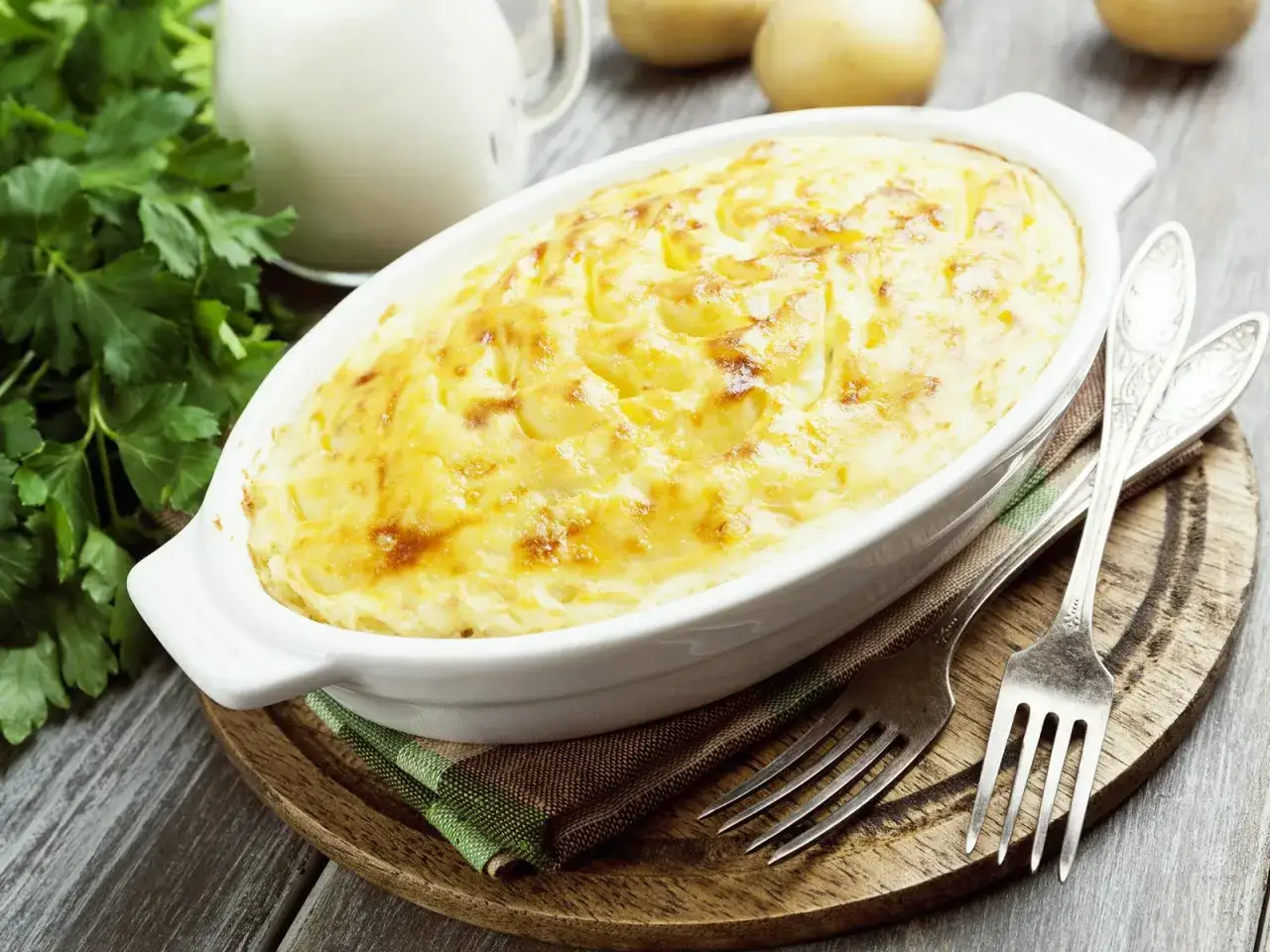 Goldbraunes, &uuml;berbackenes Kartoffelgratin ohne Sahne in einer wei&szlig;en Auflaufform, daneben Petersilie und Gabeln.