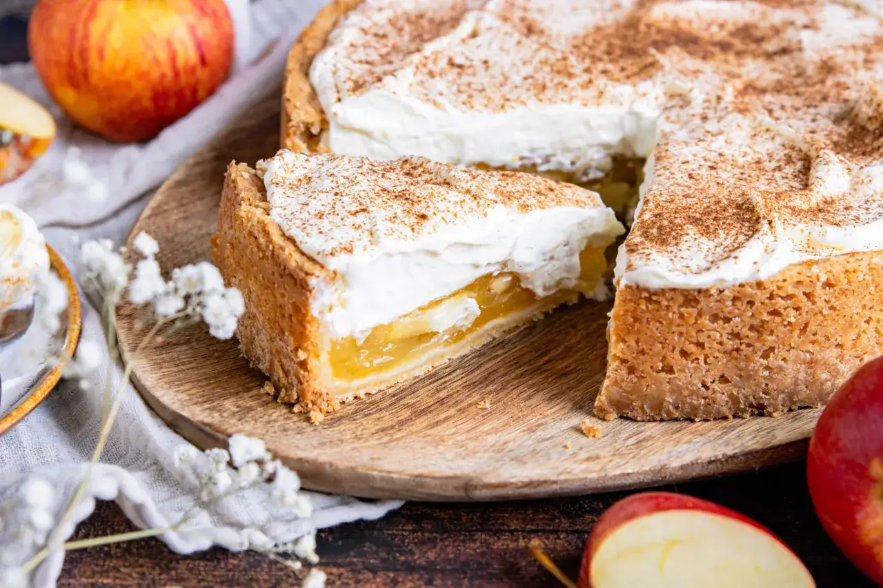 Ein Stück Apfel Schmand Torte mit saftigen Äpfeln und luftiger Sahne, bestreut mit Zimt. Frische Äpfel liegen daneben.