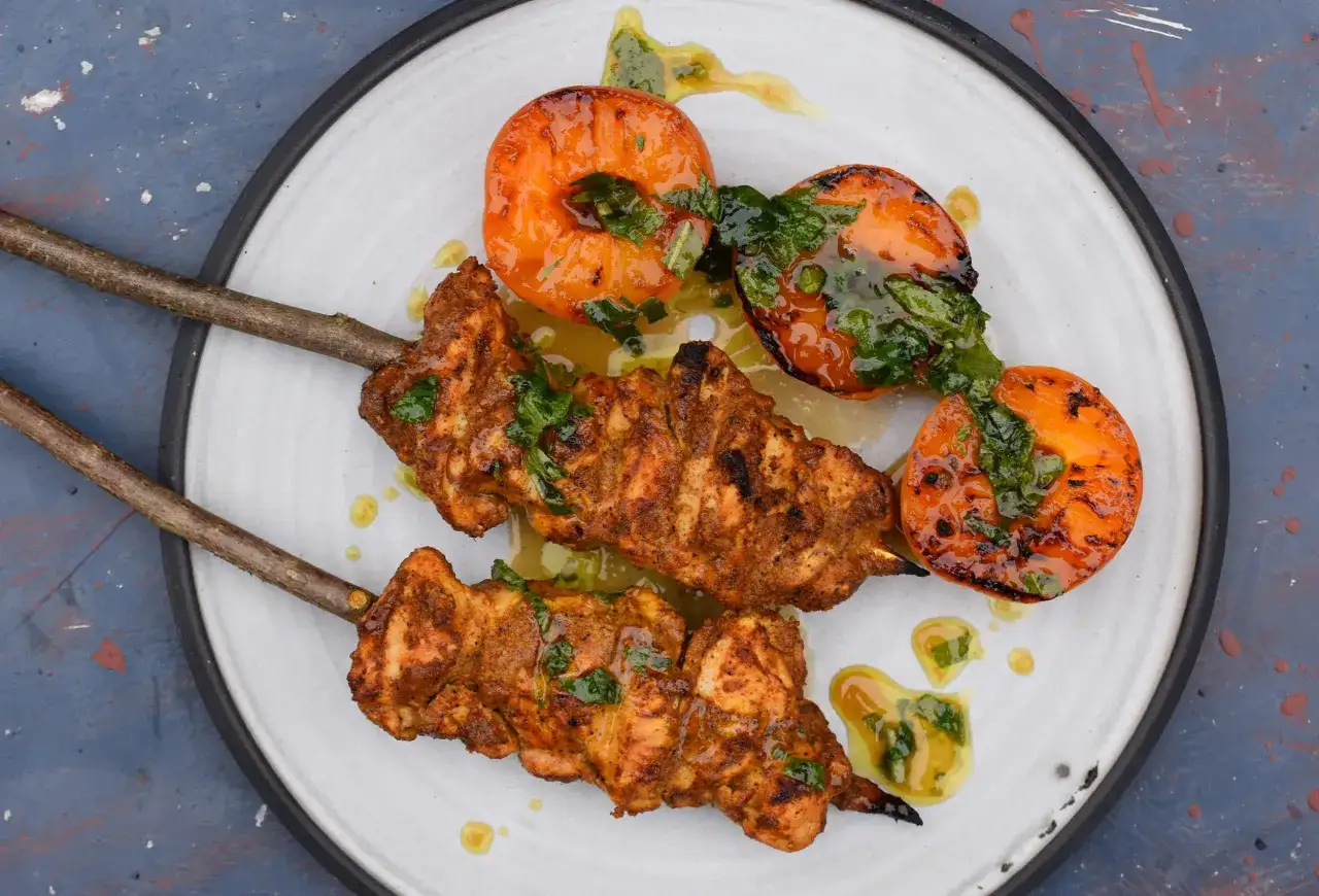 Zwei Spieße mit saftigem Tandoori Chicken, garniert mit frischen Kräutern und gegrillten Aprikosen, serviert auf einem Teller.