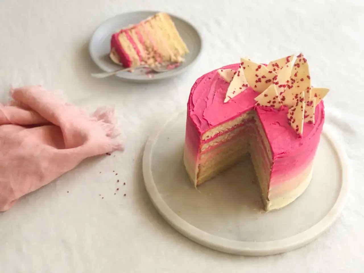 Eine mehrschichtige Buttercreme Torte Geburtstag mit pinker Glasur und wei&szlig;en Schokoladenst&uuml;cken. Ein St&uuml;ck ist bereits angeschnitten.