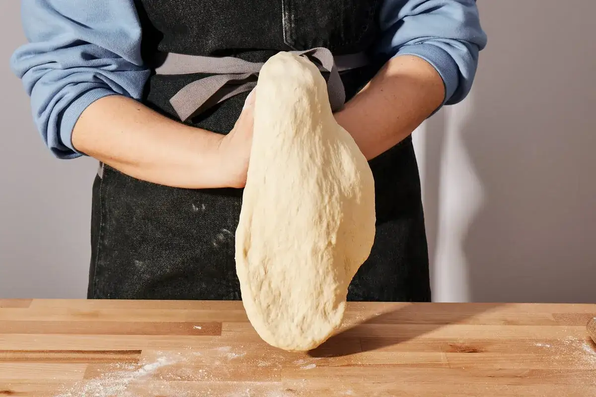 Hände formen den weltbesten Pizzateig mit Trockenhefe. Der Teig hängt elastisch zwischen den Händen, bereit für die Pizza.