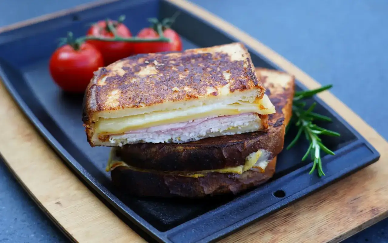 Ein goldbrauner Monte Cristo Sandwich mit Schinken und Käse, serviert mit Tomaten und Rosmarin auf einer Gusseisenplatte.