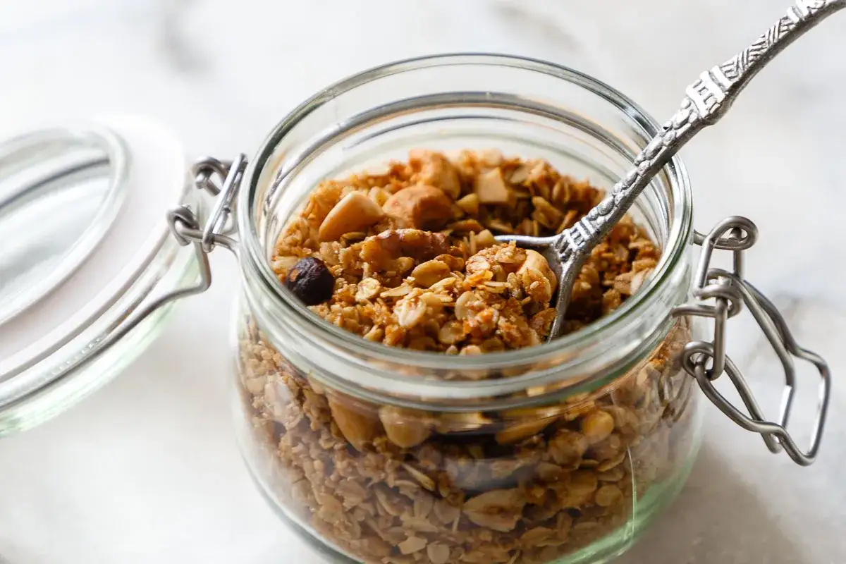 Glas gefüllt mit selbstgemachtem Granola, Nüssen und Haferflocken. Eine Löffelspitze ragt heraus.