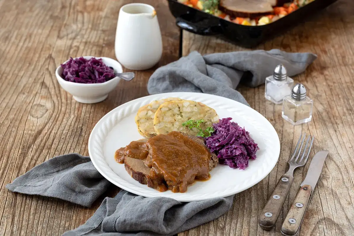 Rinderbraten aus dem Dutch Oven, serviert mit Kl&ouml;&szlig;en und Rotkohl auf einem Holztisch.