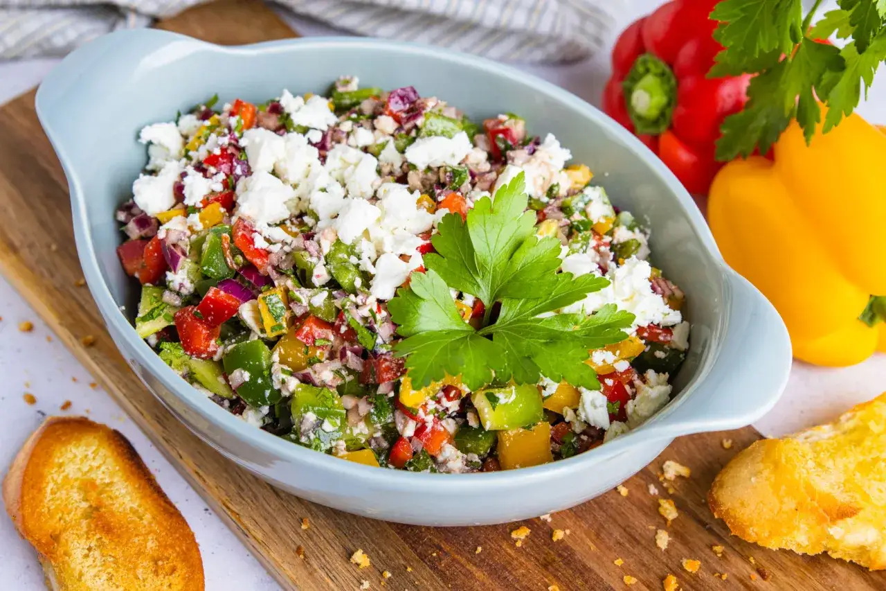Bunter Paprikasalat mit Feta, garniert mit Petersilie, serviert in einer blauen Sch&uuml;ssel neben ger&ouml;stetem Brot und ganzen Paprika.