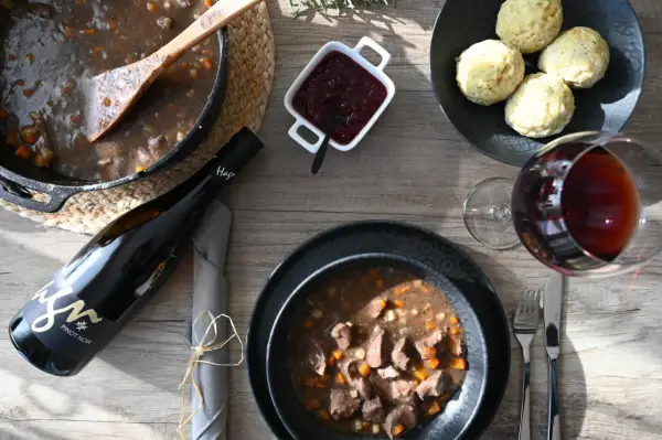 Herzhaftes Wild im Dutch Oven, serviert mit Klößen, Preiselbeeren und einem Glas Rotwein. Ein Festmahl!