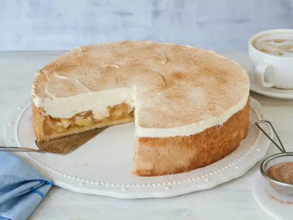 Saftige Apfel-Schmand-Torte mit Zimt bestäubt, ein Stück herausgeschnitten, daneben eine Tasse Cappuccino.