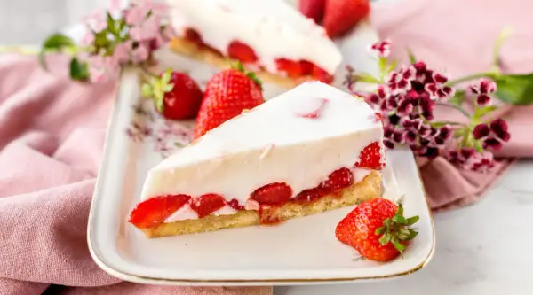 Ein Stück cremiger Erdbeerkuchen, der an Omas beste Kuchen Rezepte erinnert, mit frischen Erdbeeren und Blumen dekoriert.