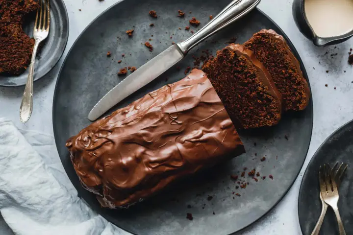 Saftiger Zucchini Schoko Kuchen mit Schokoladenglasur, angeschnitten auf einem dunklen Teller, daneben eine Gabel und ein Messer.