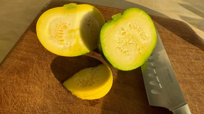 Zwei Hälften und ein Stück gelbe Zucchini liegen auf einem Holzbrett neben einem Messer.